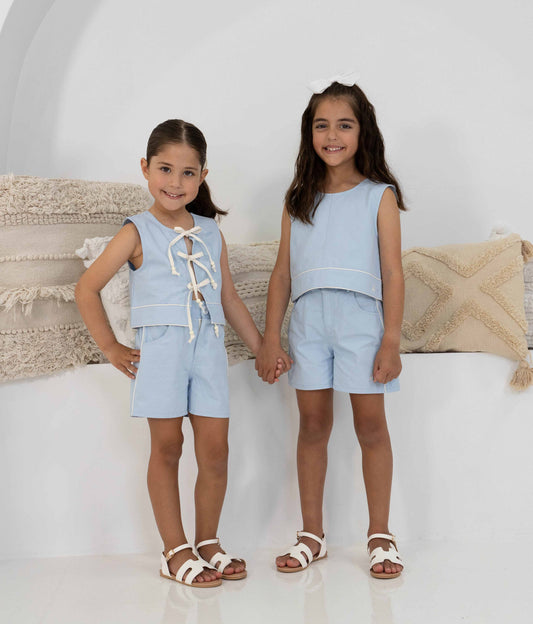 Two girls wearing matching Alice Sets, in light blue cotton two-piece sets, consisting of lace-up front top and shorts, standing against a neutral background. Displayed showing the top worn 2ways.