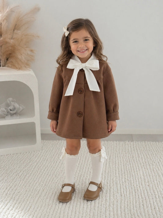 Young girl wearing a brown coat with a white bow and white knee high socks standing in a room.