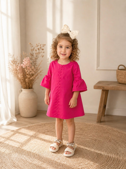 Toddler girl in a pink dress standing in a sunlit room with a wooden bench and vase in the background.