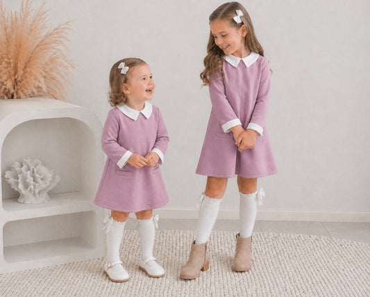 Two young girls in the Ariella winter dress paired with knee high white socks, one is wearing white mary jane shoes and the other in cream ankle boots. They're smiling, standing on white carpet.
