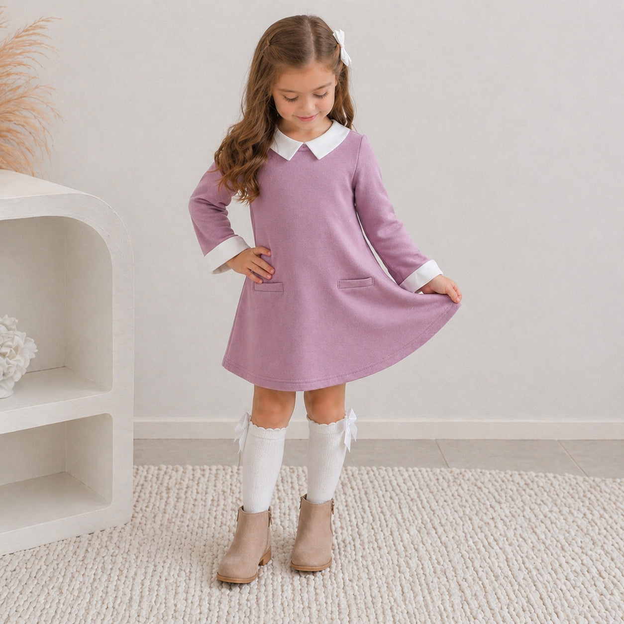 7 yr old girl in the Ariella winter dress paired with knee high white socks with cream ankle boots. She's posing looking down, holding the corner of the dress and smiling, standing on white carpet.