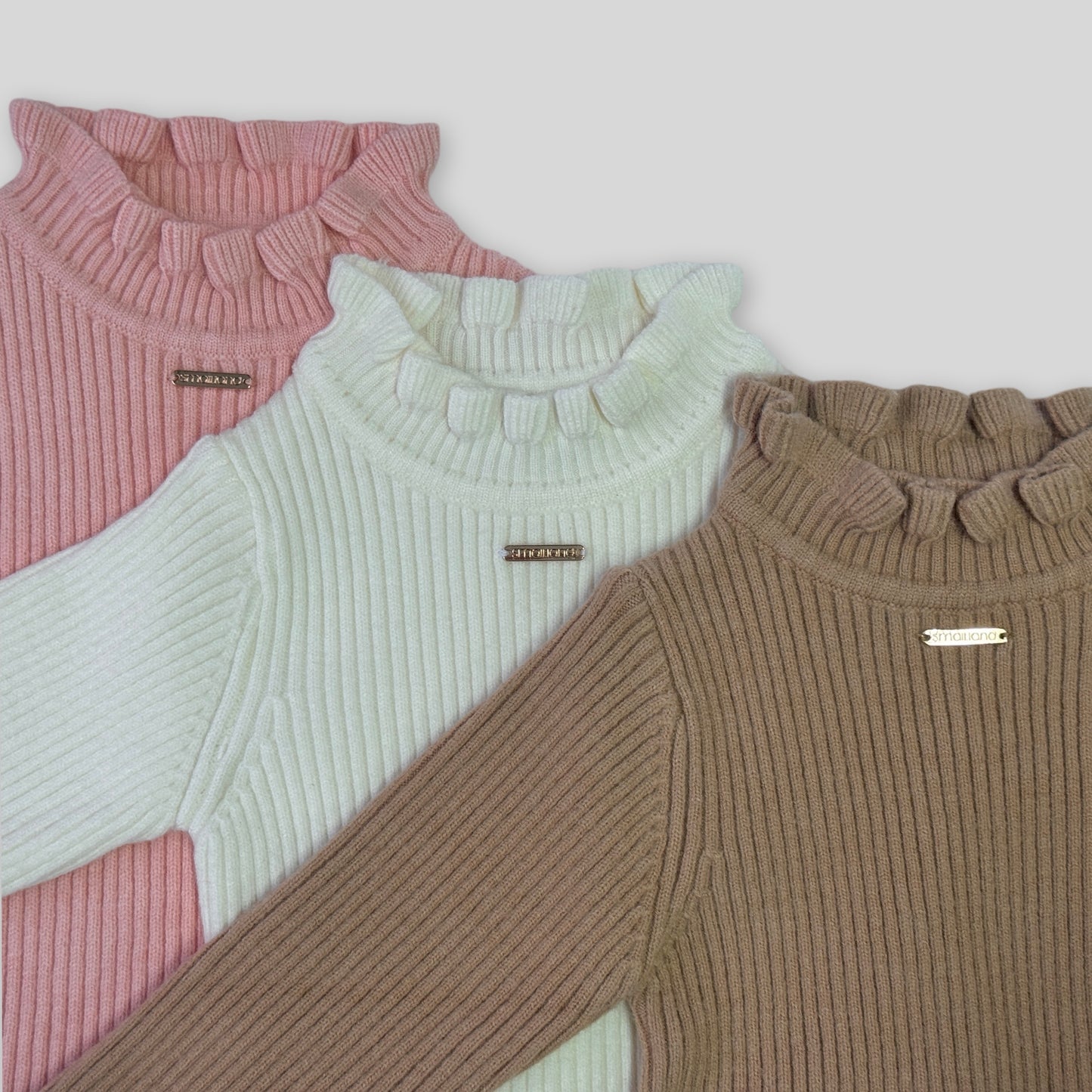 Close up photo of the gigi girls winter long sleeve knit showing fabric and details, with a rose gold metal tab logo at the centre front chest. Also shows the frill turtle neck design 