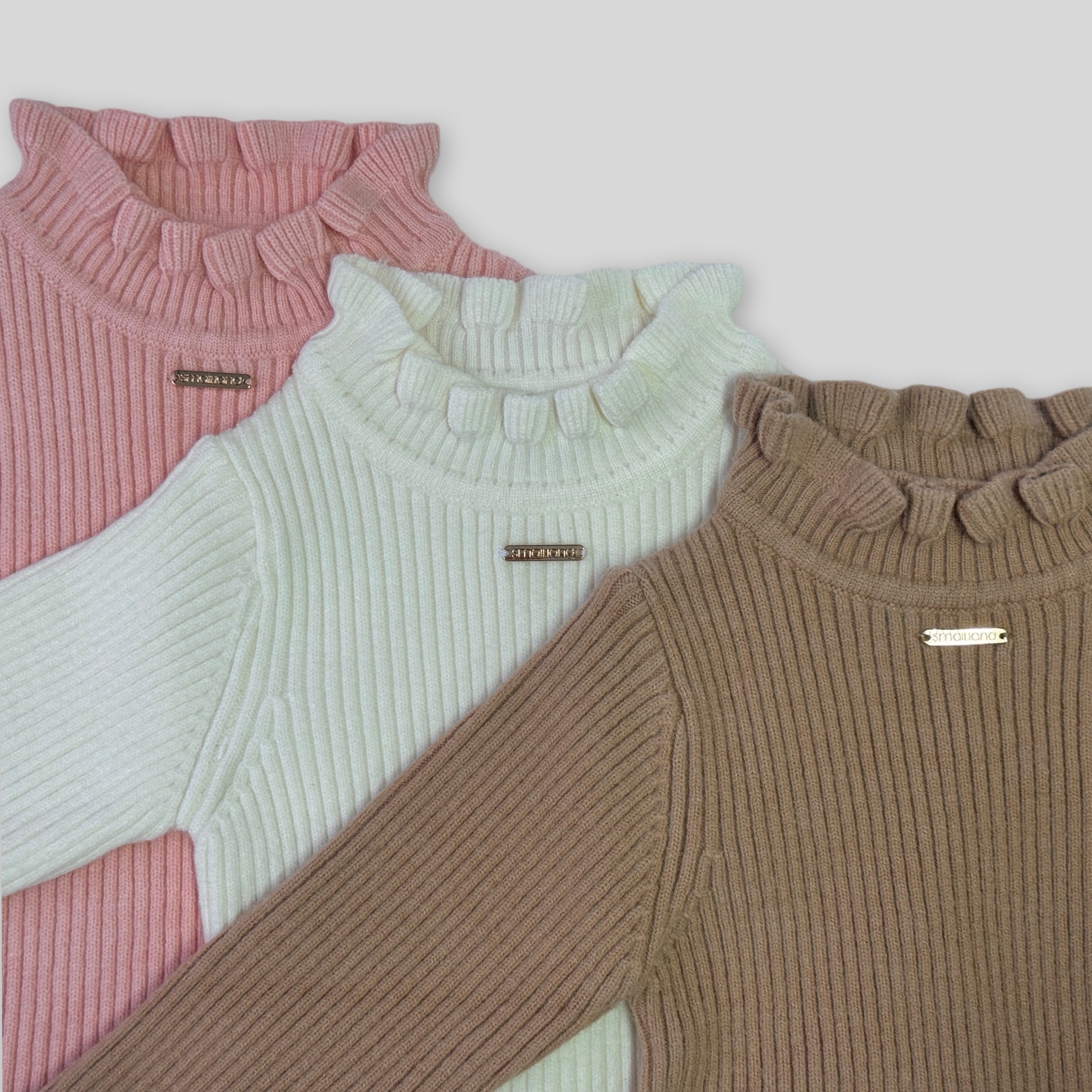 Close up photo of the gigi girls winter long sleeve knit showing fabric and details, with a rose gold metal tab logo at the centre front chest. Also shows the frill turtle neck design 