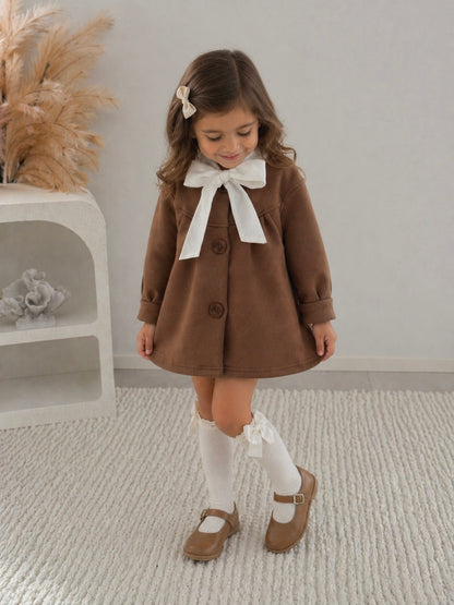 Young 5yr old girl in a brown dress with a white bow and white knee high socks paired with brown mary jane shoes, looking down, smiling standing in a room with a neutral background.
