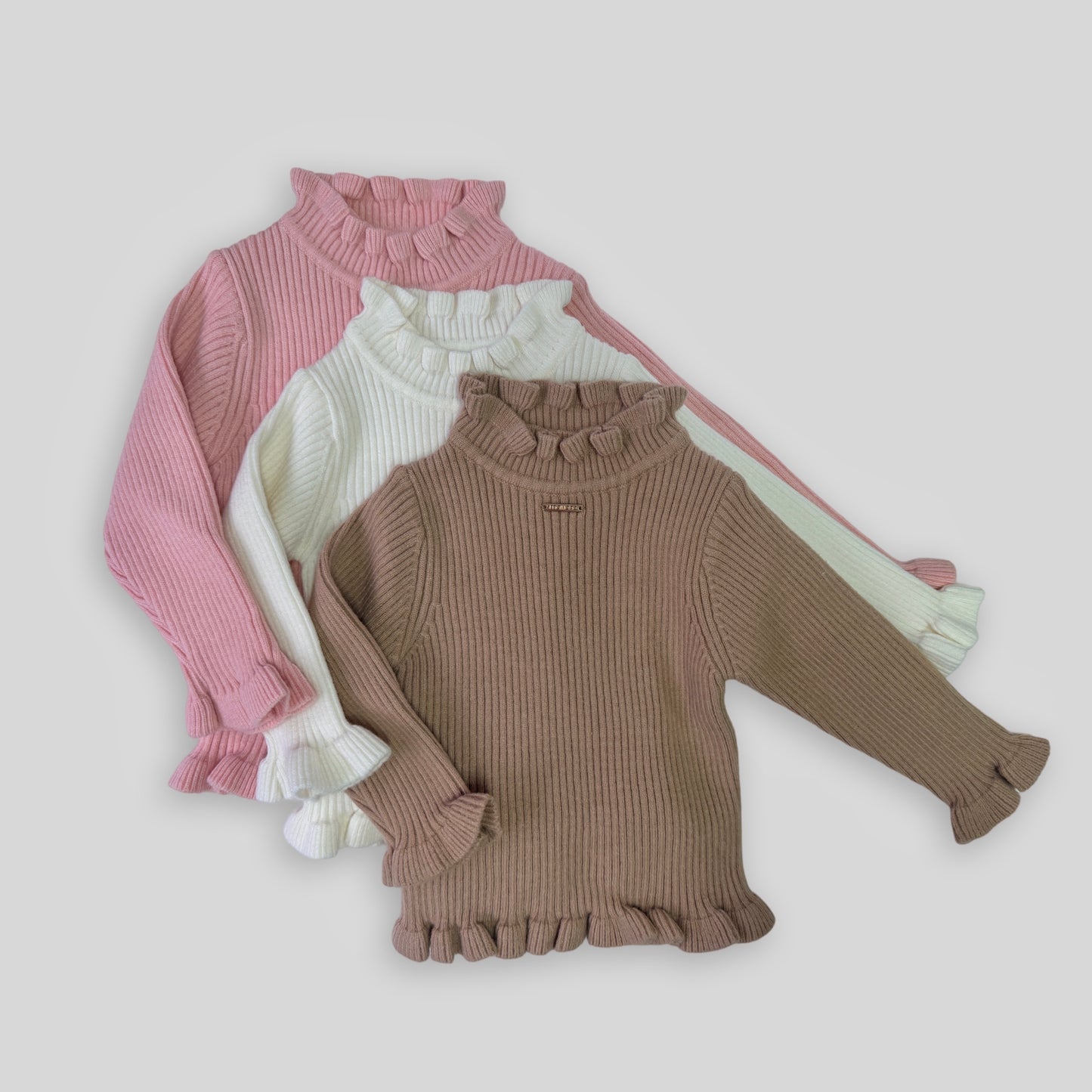 A flat-lay image of the gigi girls winter long sleeve knit showing fabric and details, with a rose gold metal tab logo at the centre front chest. Also shows the frill turtle neck design, frills on the cuff and hem line of the winter knit