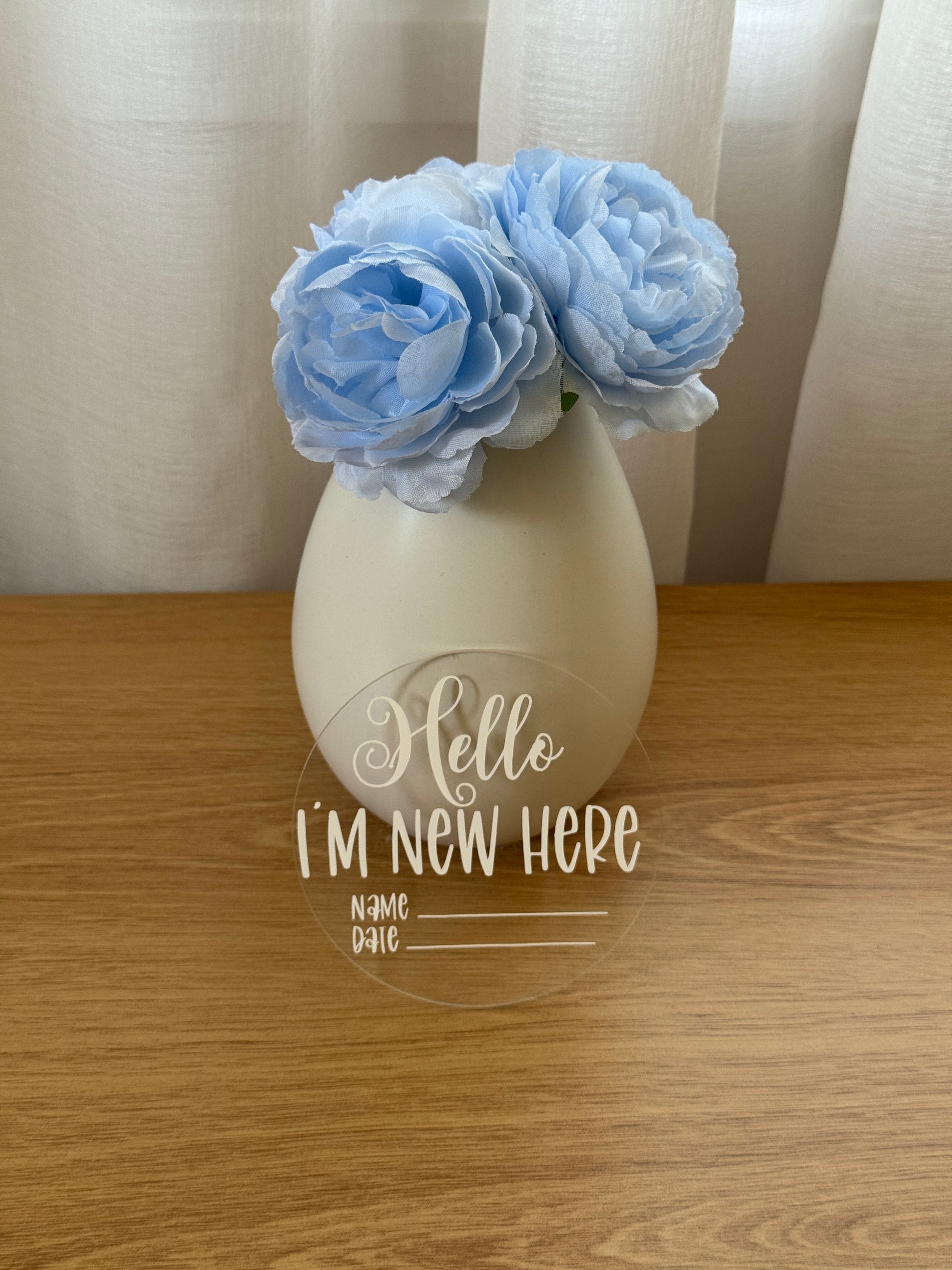 Decorative sticker with 'Hello, I'm New Here' text on a acrylic clear disk leaning on a vase with blue flowers against a wooden background