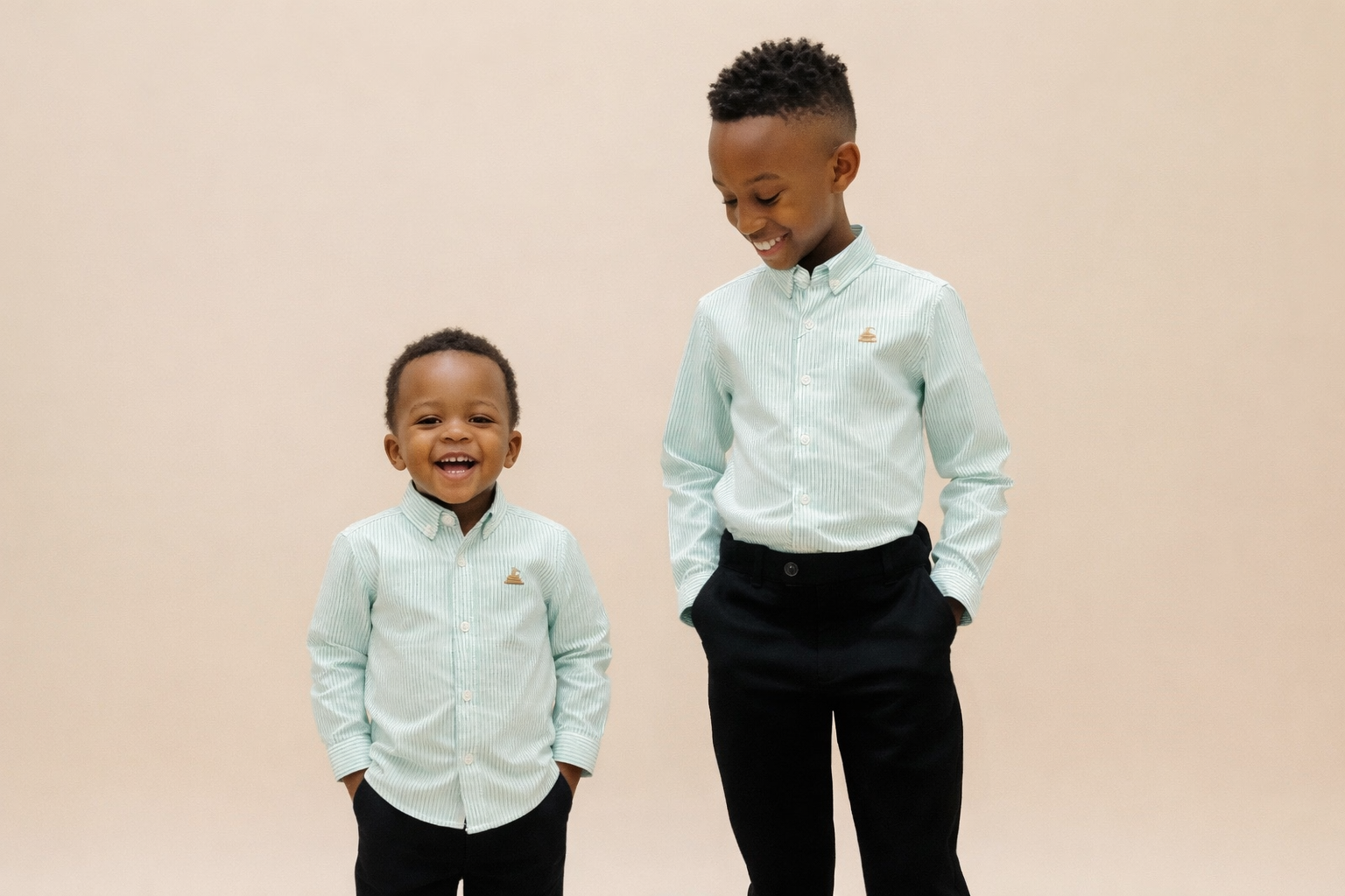 Two young boys wearing light green button-up shirts and black pants on a beige background