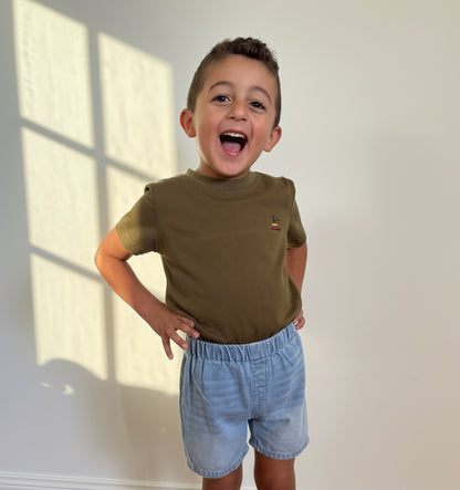 Child wearing a green shirt and denim shorts standing on a wooden floor.