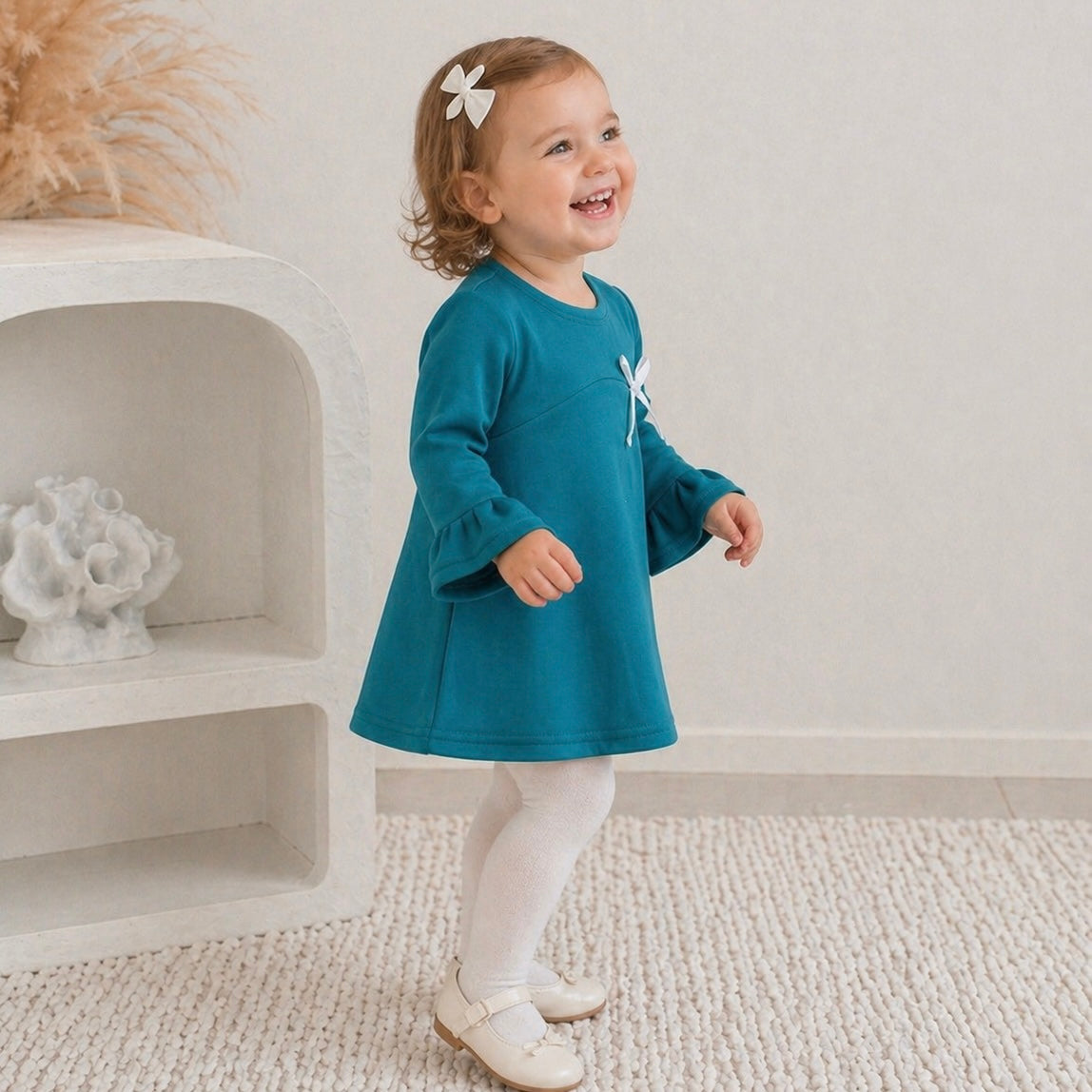 3yr old toddler girl wearing the Maya mini winter dress in teal green, smiling, standing on some white carpet.