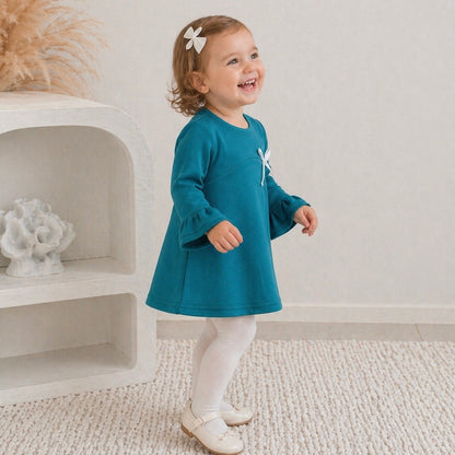 3yr old toddler girl wearing the Maya mini winter dress in teal green, smiling, standing on some white carpet.