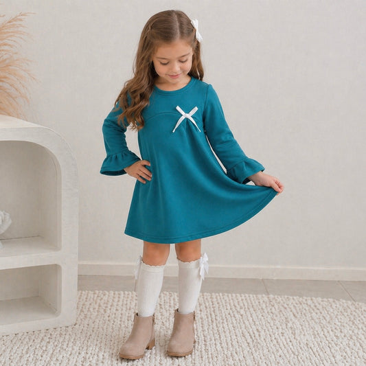 7yr old toddler girl wearing the Maya mini winter dress in teal green, looking down and posing, standing on some white carpet.
