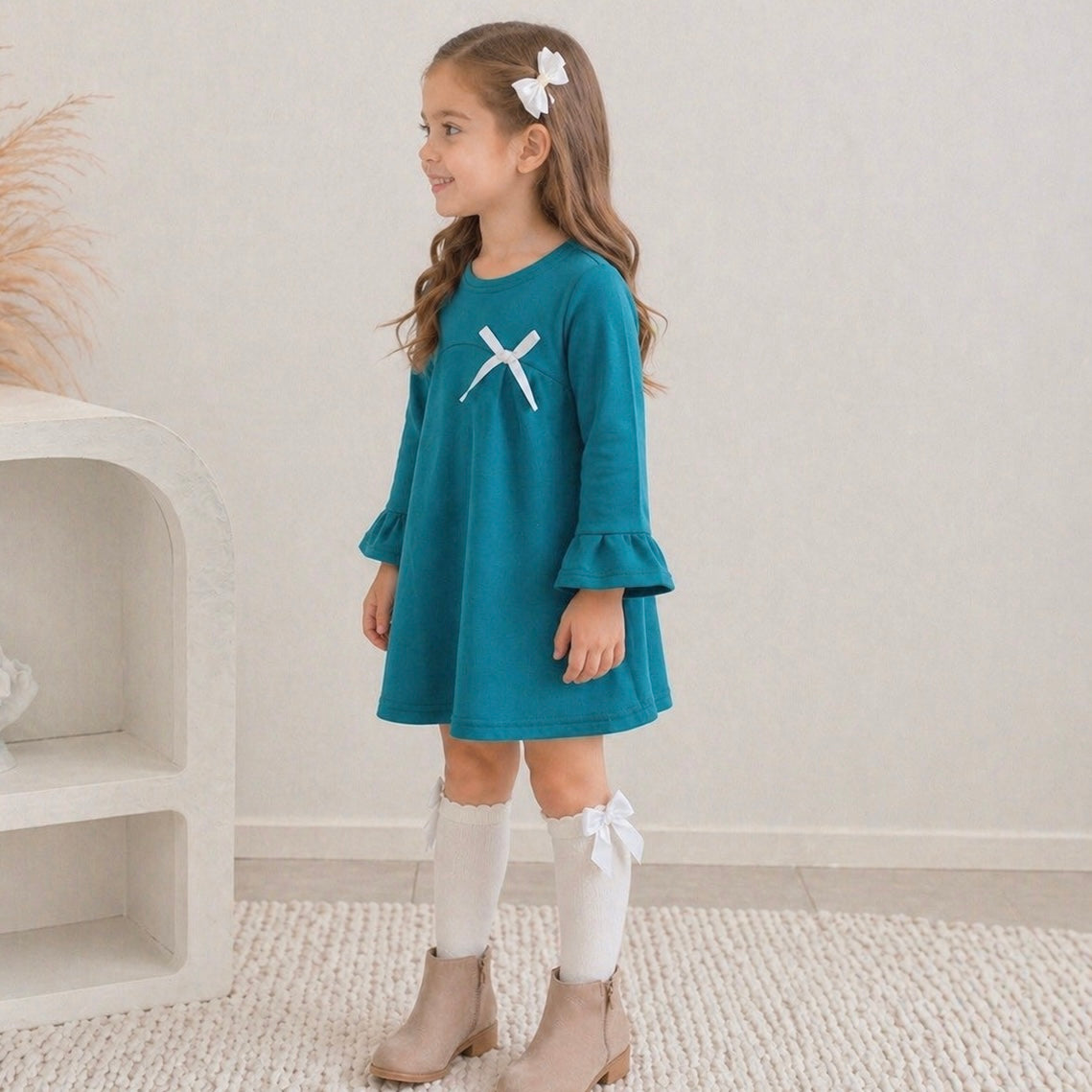 7yr old toddler girl wearing the Maya mini winter dress in teal green, looking left smiling, standing on some white carpet.
