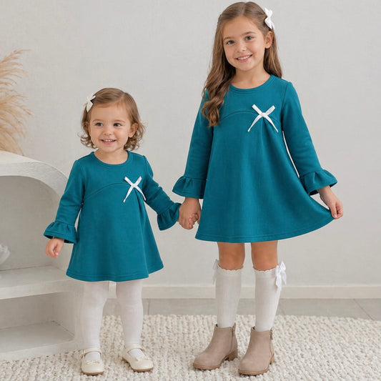 Two young toddler girls wearing the Maya mini winter dress in teal green, standing on some white carpet.