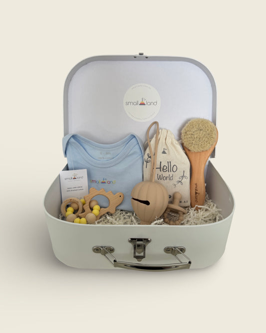 A blue suitcase-style gift box with various baby items like a onesie, milestone blocks, a hairbrush, a teething ring, and a dummy case holder, all presented in a stylish manner.