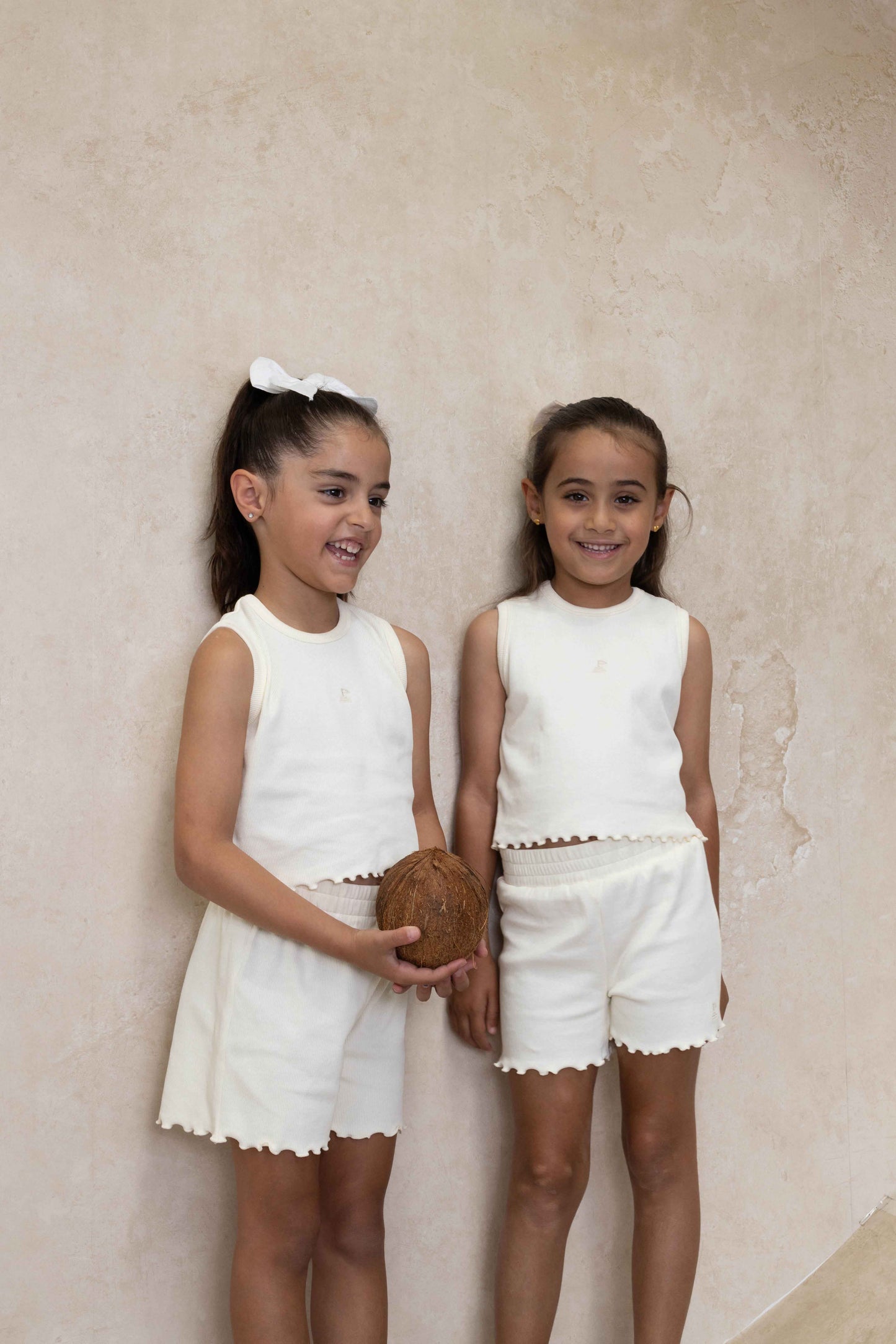 Two girls wearing matching cream ribbed tank tops and shorts with lettuce-edge hem, standing against a plain wall.