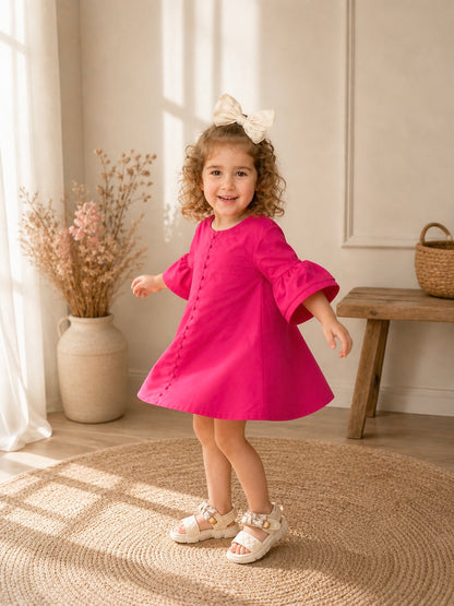 Toddler girl in a pink dress about to twirl in a sunlit room with a wooden bench and vase in the background.