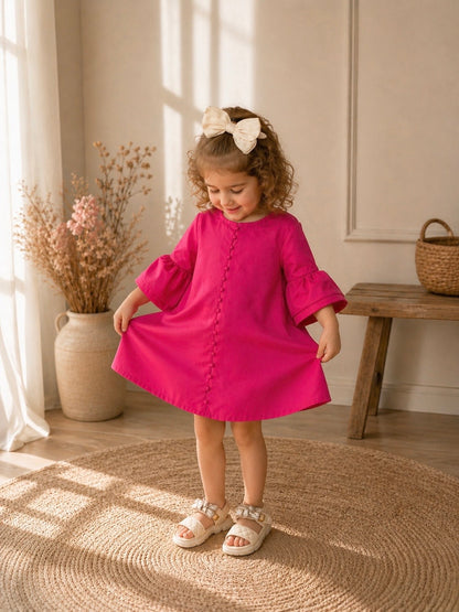 Toddler girl in a pink dress looking down holding the dress, standing, smiling in a sunlit room with a wooden bench and vase in the background.
