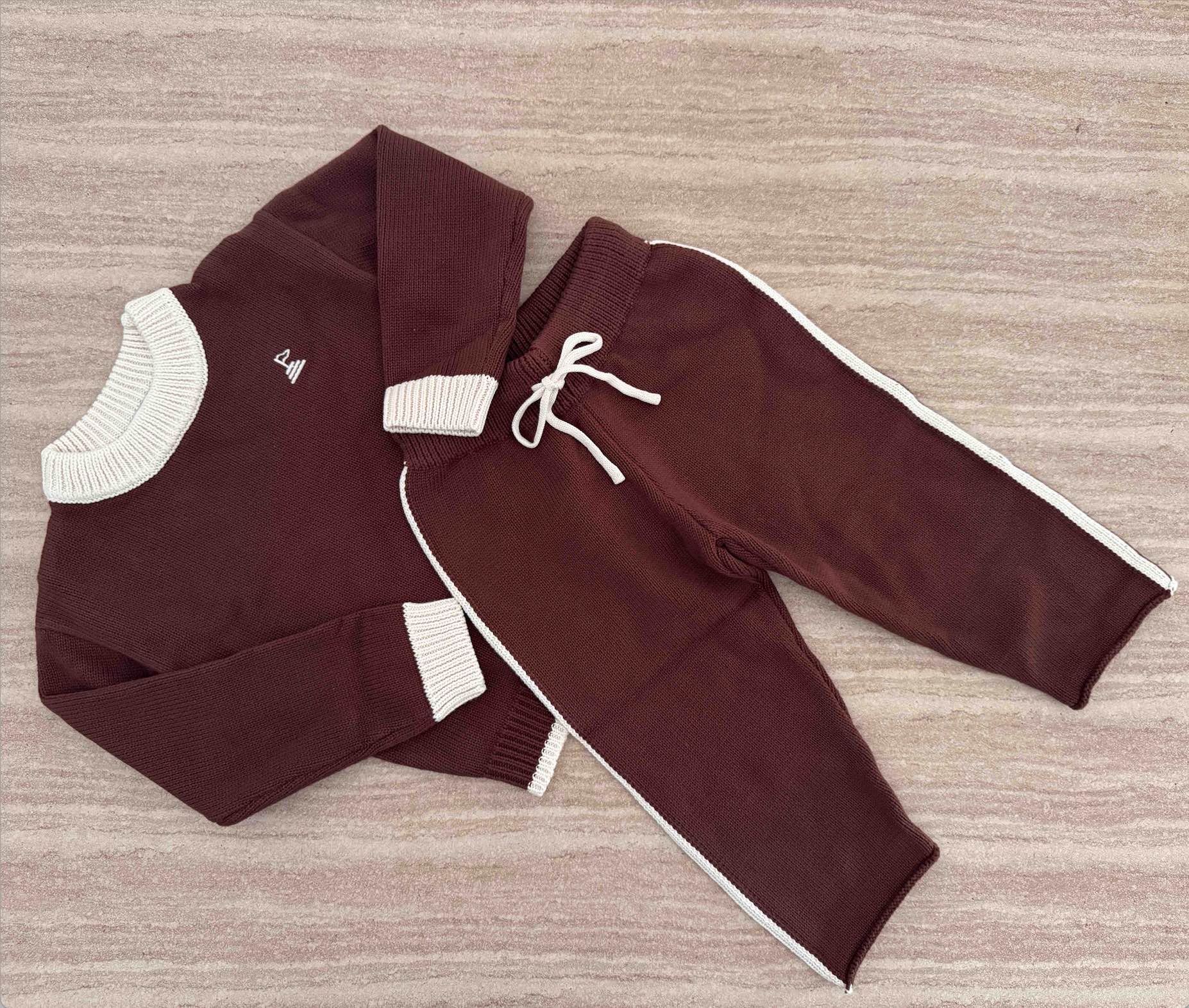 A two-piece children's knit outfit laid flat on a textured surface, featuring a long-sleeve sweater and matching jogger pants in deep brown with cream ribbed trims/ drawstring.
