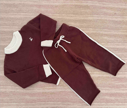 A two-piece children's knit outfit laid flat on a textured surface, featuring a long-sleeve sweater and matching jogger pants in deep brown with cream ribbed trims/ drawstring.