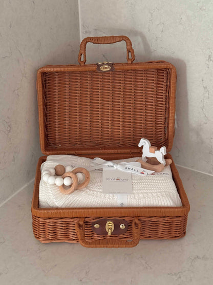 A brown woven wicker suitcase opened to reveal a baby gift set inside, including a white knit blanket, wooden teething ring, and a white wooden rocking horse, set against a light gray marble background.