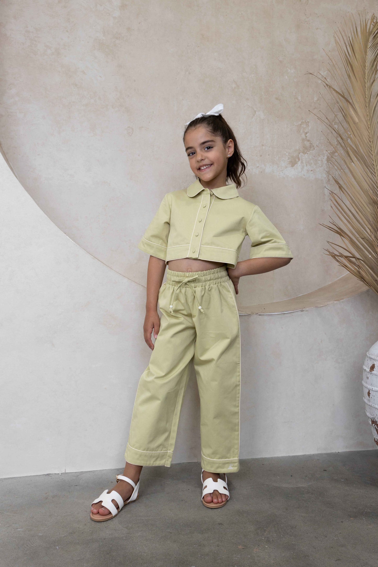 A girl model wearing high-waisted, relaxed-fit light shadow green pants with contrast white piping on side seams and an adjustable drawstring waist.