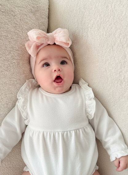 Baby wearing a white outfit and pink headband with a bow, lying on a beige surface.