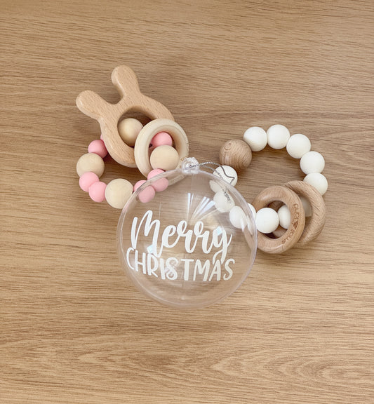 Wooden teething ring with colorful beads and a clear ornament with 'Merry Christmas' text on a wooden surface.
