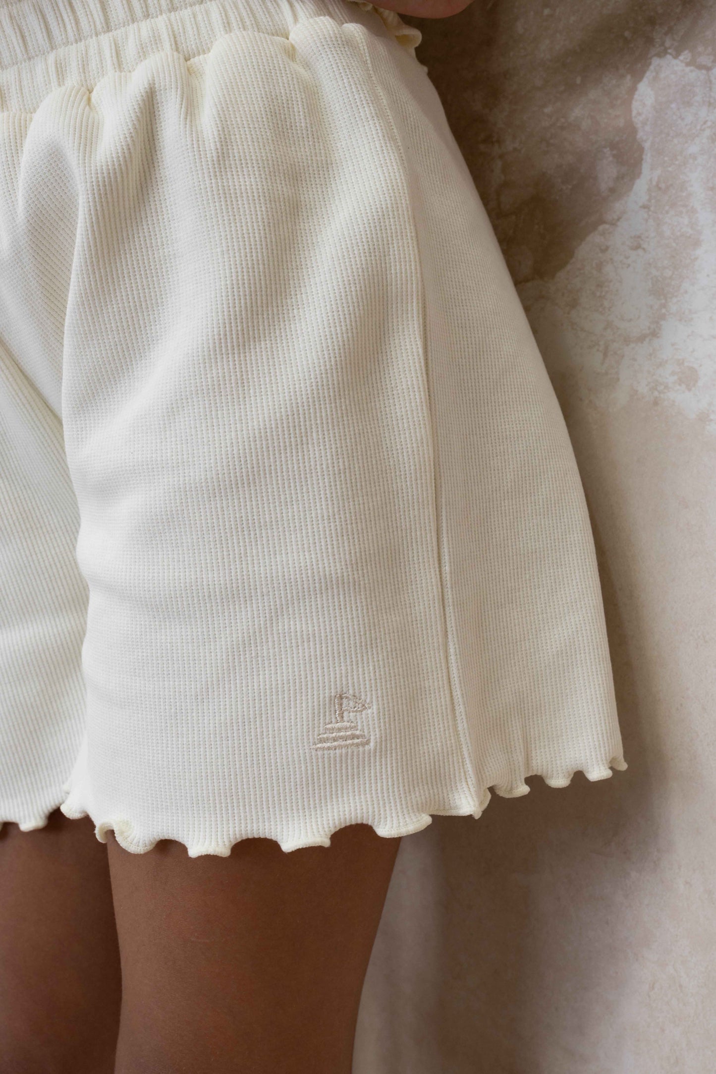 close up photo of cream ribbed shorts with logo embroidered at side seam, and lettuce edge frills