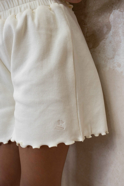 close up photo of cream ribbed shorts with logo embroidered at side seam, and lettuce edge frills