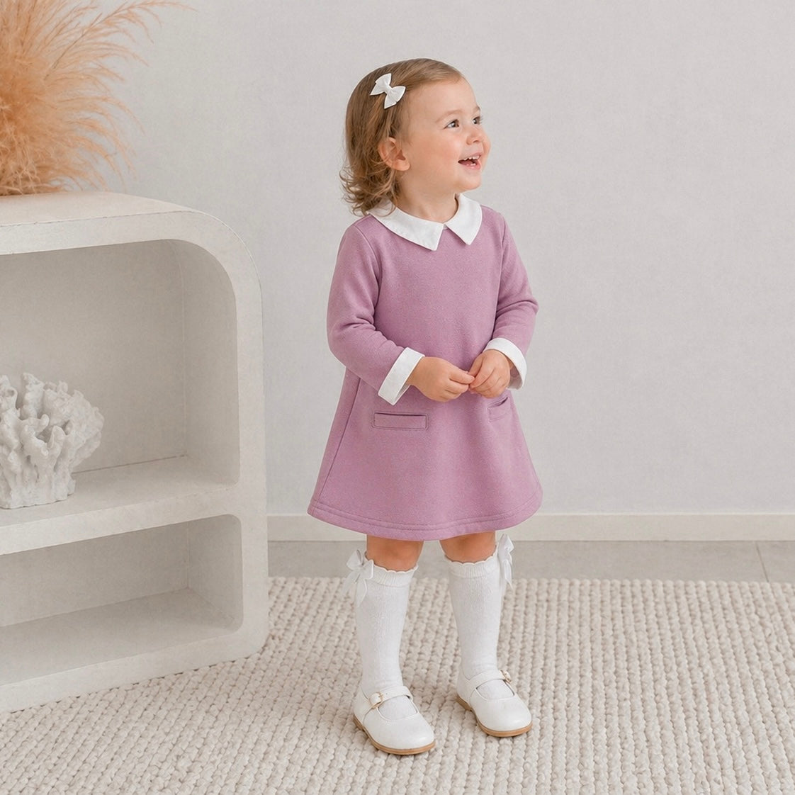  3yr old girl in the Ariella winter dress paired with knee high white socks with white mary jane shoes. She's looking up smiling, standing on white carpet.