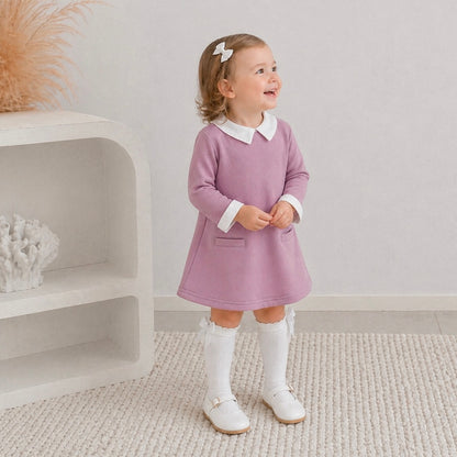  3yr old girl in the Ariella winter dress paired with knee high white socks with white mary jane shoes. She's looking up smiling, standing on white carpet.