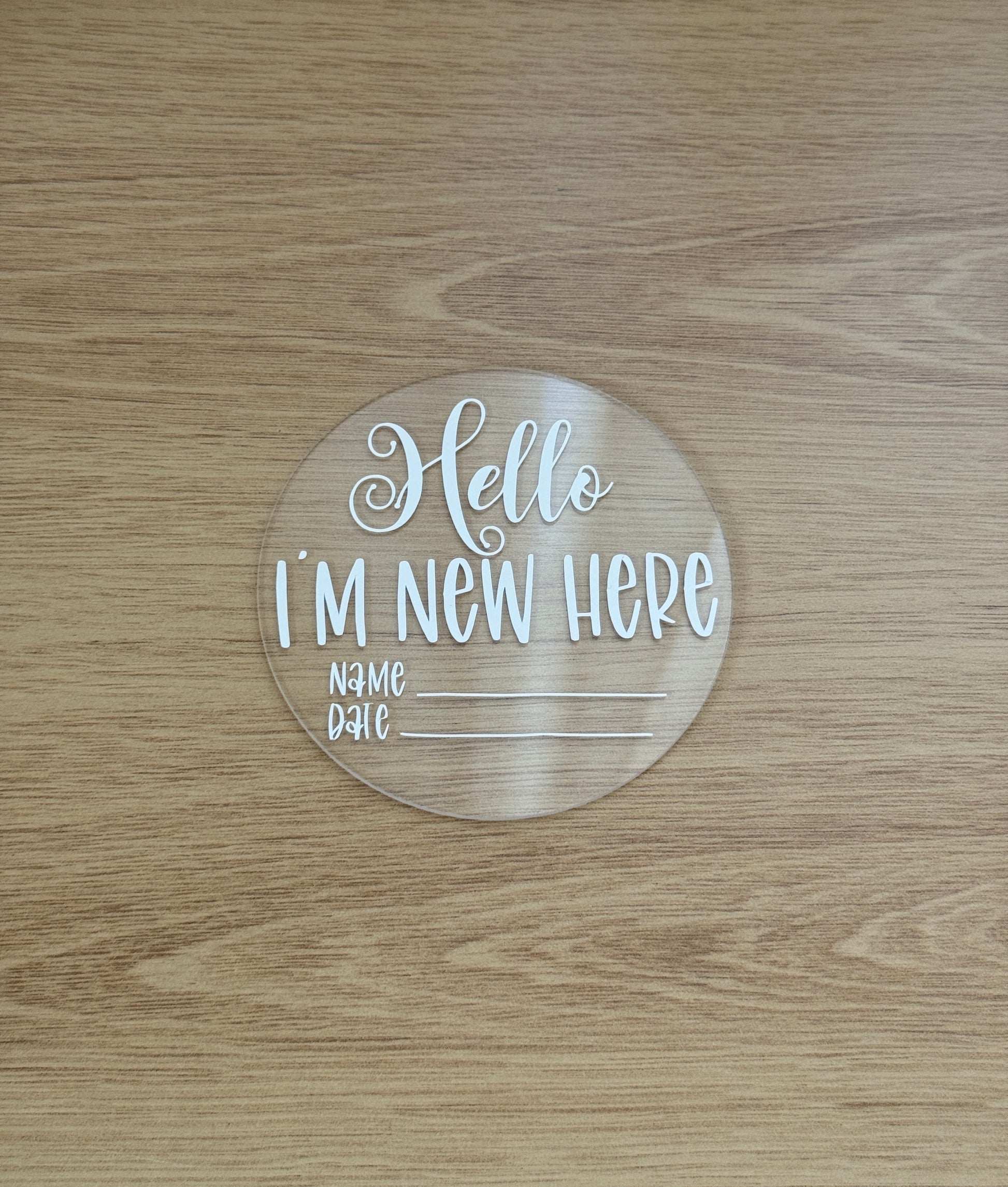 Round acrylic sign with 'Hello I'm New Here' text on a wooden surface