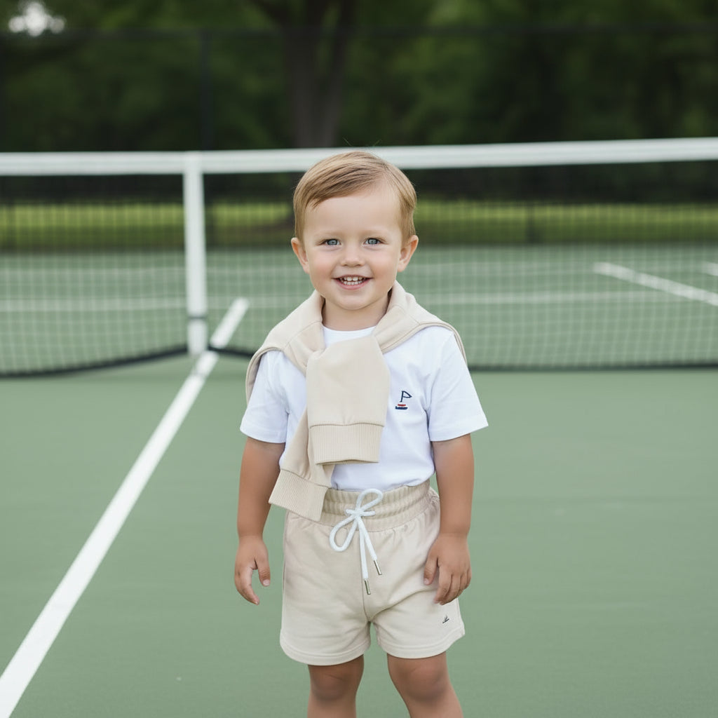Toddler boy standing on a tennis court with our signature unisex biscotti colour sweatshirt over their shoulder, wearing small land signature white tee and our signature unisex biscotti colour kids shorts